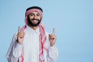sonriente musulmán hombre vistiendo tradicional cultura ropa y señalando arriba a Copiar espacio con alegre expresión retrato. despreocupado árabe demostración hacia arriba para anuncio y mirando a cámara foto