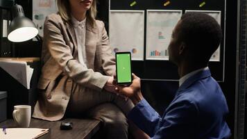 African american detective conducts investigation with cellphone displaying greenscreen, collaborating with policewoman. Male inspector vertically grasps mobile phone with blank chromakey template. photo