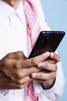 Muslim man holding mobile phone in arms, sending online message while using chatting app closeup. Arab in traditional thobe using smartphone and scrolling social media page photo