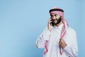 Furious muslim man wearing traditional clothes getting mad while talking on mobile phone. Aggressive arab in thobe standing with clenched fist and angry expression while answering smartphone call photo