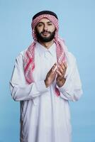 confidente árabe hombre en tradicional túnica aplausos manos en aplausos mientras en pie en estudio. musulmán persona vistiendo islámico atuendo aplaudiendo, demostración apoyo y mirando a cámara foto