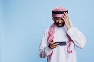 Disappointed arab loosing mobile game competition while enjoying digital entertainment in studio. Unhappy unsuccessful muslim man playing videogame on smartphone with upset expression photo