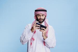 Muslim man wearing traditional clothes playing online game on smartphone while posing in studio. Arab gamer in thobe and islamic headscarf enjoying entertainment app on mobile phone photo