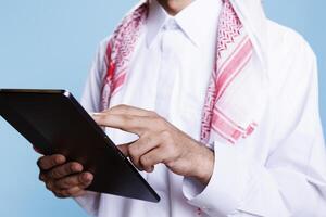 Man dressed in traditional muslim clothes holding digital tablet in hands and tapping on screen closeup. Person wearing thobe using portable gadget and browsing internet page photo