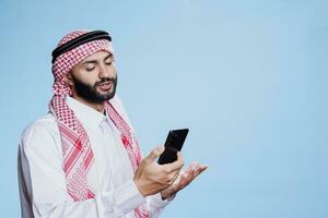 Muslim man dressed in traditional attire engaging in video chat on smartphone, connecting remotely. Arab in ghutra and thobe having conversation in online meeting using mobile phone photo