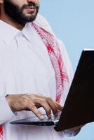 Man wearing traditional muslim clothes typing on laptop keyboard closeup. Arab person hand writing online message while standing with portable computer in studio close view photo