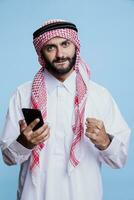 Excited muslim man wearing traditional clothes using smartphone, showing yes gesture with clenched fist portrait. Arab dressed in thobe and ghutra holding mobile phone and posing with winner concept photo