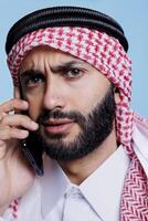 Arab man frowning eyebrows and looking at camera with confused expression while chatting on smartphone. Muslim person wearing ghutra speaking on mobile phone closeup portrait photo