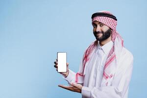 Smiling muslim man in traditional thobe presenting smartphone with empty white screen portrait. Confident arab showcasing mobile phone blank touchscreen and looking at camera photo