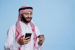 Smiling muslim man using debit card to pay online while shopping in internet store with smartphone. Arab in islamic thobe and headscarf making purchase on mobile phone website photo