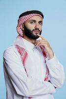 Thoughtful muslim man wearing thobe and checkered headscarf standing in deep thoughts with pensive emotion. Questioned arab person posing in contemplation on blue background photo