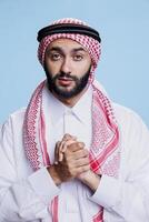 Muslim man praying while showing faith and belief and looking at camera with hopeful expression. Arab prayer posing with folded hands while praising god studio portrait on blue background photo