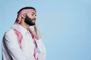 Thoughtful muslim man wearing traditional clothes touching chin while posing in studio with pensive expression. Puzzled arab person in thobe and headdress thinking about solution photo