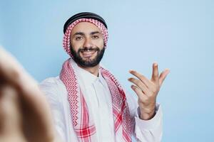 Cheerful muslim man wearing traditional clothes engaging in video call from pov perspective. Smiling arab person in thobe and headscarf looking at front camera while speaking in online meeting photo