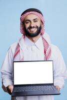 Smiling muslim man showing laptop with blank screen mockup for software advertising portrait. Arab presenting computer with empty display and looking at camera with cheerful expression photo