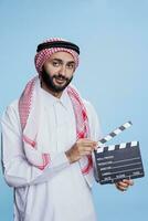Muslim man in traditional clothes posing with movie slate and looking at camera with smiling expression. Arab person showcasing film production clapperboard studio portrait photo