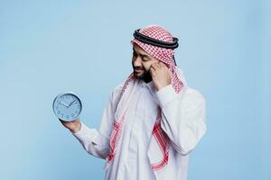 Man wearing traditional muslim clothes holding retro alarm clock and looking at time. Arab person dressed in thobe and headscarf showing punctuality and schedule deadline countdown concept photo