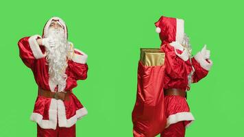 Santa does thumbs up and thumbs down against greenscreen background, carrying sack of presents on camera. Undecided saint nick shows like and dislike sign, wearing famous costume. photo