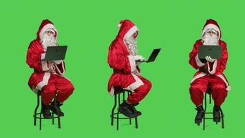 Modern santa character on laptop in full body greenscreen studio, sitting on chair and browsing social media app on wireless pc. Young cool saint nick cosplay in red festive costume. photo