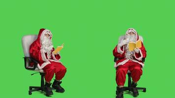 Wise person with lecture hobby sitting on chair and reading fairytales or literature in studio. Santa claus holding fiction or poetry book, sits over full body greenscreen backdrop. photo