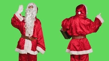 Man in santa claus suit waving hello and hitchhiking with suitcase, carrying baggage while he poses over greenscreen backdrop. Cheerful festive person acting positive on christmas eve. photo