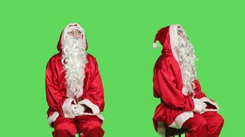 Man dressed as santa claus on chair sitting against greenscreen background, father christmas embodiment. Young adult advertising december seasonal holiday, traditional event. photo