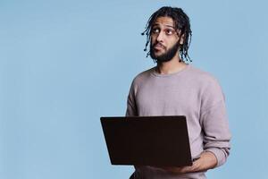 Pensive arab man thinking about business report while holding laptop and looking up. Thoughtful person with puzzled facial expression analyzing start up project on portable computer photo