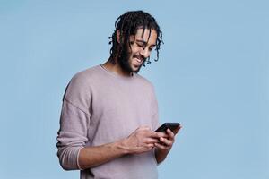 Smiling arab man texting on smartphone with happy face expression. Cheerful young person in casual clothes chatting in social media, sending online message on mobile phone photo