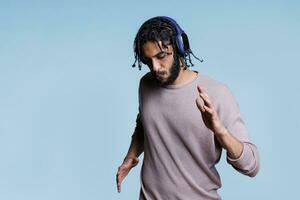 concentrado árabe hombre bailando mientras escuchando a música en auriculares. joven persona enfocado en canciones lista de reproducción en inalámbrico auriculares Moviente manos y cuerpo en azul antecedentes foto