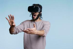 Excited man in vr glasses exploring virtual reality and touching objects in metaverse. Person using wearable headset and playing video game while making gestures with hands photo