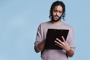 Concentrated arab writing on clipboard, filling application form with focused facial expression. Young thoughtful person standing while taking notes, reading and checking list photo