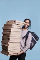 Carefree arab deliveryman carrying big pizza boxes pile and looking at camera with cheerful expression. Smiling restaurant delivery service courier standing huge stack of fast food packages photo