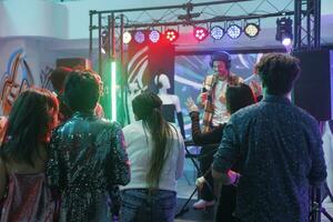 personas bailando y ir de fiesta en Club nocturno mientras joven hombre DJ ejecutando en etapa iluminado con focos multitud clubbing y Moviente en pista de baile a electrónico música concierto foto
