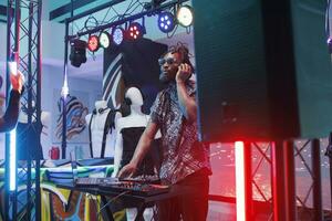 Dj in headphones mixing sounds using digital electronic music controller on stage in nightclub. Young african american man musician performing at discotheque party in club photo