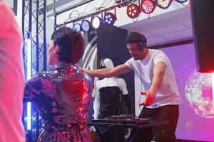 joven hombre DJ mezcla techno pistas en etapa mientras personas ir de fiesta en pista de baile en Club nocturno. músico jugando electrónico música en etapa mientras clubbers bailando a discoteca foto