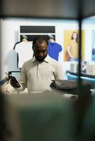 africano americano hombre comprobación vestir precio en en línea sitio web en teléfono inteligente mientras elegir atuendo en ropa almacenar. cliente utilizando móvil teléfono mientras seleccionando prenda en centro comercial foto