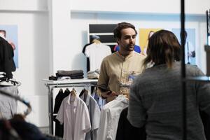 Mall worker standing near clothes rack and checking apparel available for purchase using digital tablet. Shopping center young man employee checking store app on portable gadget photo