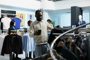 African american man shopping for outfit in clothing store and checking size options in smartphone app. Mall client searching for garment information on mobile phone website photo