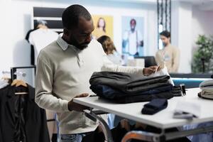 African american man shopping for apparel in mall using smartphone to access clothing store mobile website. Customer selecting shirt from hanging rack and scrolling available stock in app photo