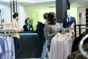 Clothing store smiling customer holding jacket on hanger and checking size options in smartphone app. African american woman choosing formal outfit and using mobile phone website photo