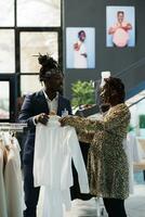 Showroom worker helping customer choosing white shirt, discussing clothes fabric in clothing store. African american pregnant woman buying fashionable maternity merchandise in modern boutique photo