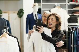 juguetón elegante mujer tomando imagen con moderno teléfono en sala de exposición, teniendo divertido durante compras sesión. alegre clientela comprobación casual vestir marcas para comercial actividad, comprando de moda ropa foto