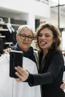 Women taking photo with phone in clothing centre, senior woman holding white shirt in front of camera. Shopaholic clients having fun in modern boutique while buying stylish clothes