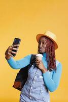 Woman taking images on holiday trip, making funny faces on mobile phone and drinking cup of coffee. Female tourist having fun with photos on camera, carrying luggage and trolley bags.