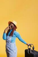 africano americano mujer con Gafas de sol dejando en fiesta con sombrero y equipaje, preparando a viaje en el extranjero. alegre persona sonriente y que lleva carretilla bolso en estudio, ocio actividad. foto