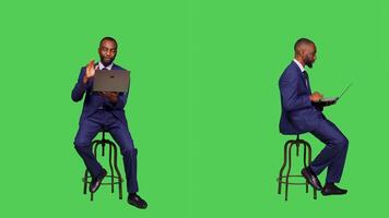 Male manager browsing website and talking on videocall, working with laptop while he sits in chair over full body greenscreen backdrop. Businessman using online conference, online network. photo