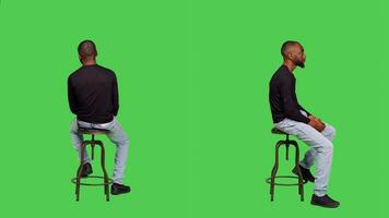 Optimistic person sitting on chair in studio, posing over full body greenscreen background. Confident male adult wearing jeans and shirt waiting for something or someone, natural man. photo