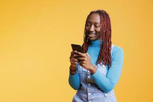 africano americano niña utilizando móvil teléfono en fiesta viaje, hojeada social medios de comunicación aplicación en estudio. joven persona comprobación en línea Internet página en teléfono inteligente, fin de semana actividades. foto