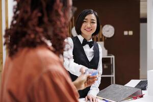 mujer pago para habitación reserva a hotel recepción, utilizando tarjeta y pos terminal para electrónico pago a frente escritorio encimera. empleado acuerdo archivos de reserva consultas a cheque afuera. foto