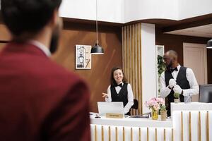 Receptionist greets hotel guest, providing excellent concierge services for businessman on work trip. Traveller with luggage being welcomed by front desk staff and bellboy, luxury resort. photo